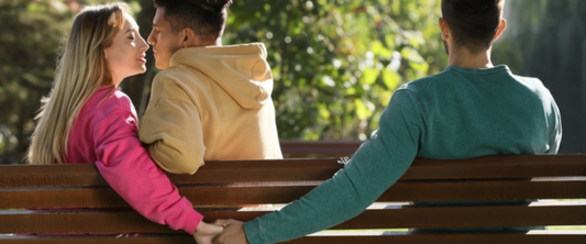 Woman holding hands with another man behind her boyfriend's back on bench in park. Love triangle