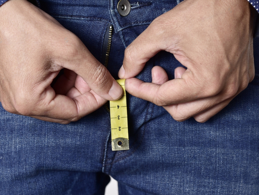 Closeup of a young man holding a short piece of measuring tape that is popping up from the fly of his jeans