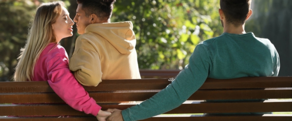 Woman holding hands with another man behind her boyfriend's back on bench in park. Love triangle
